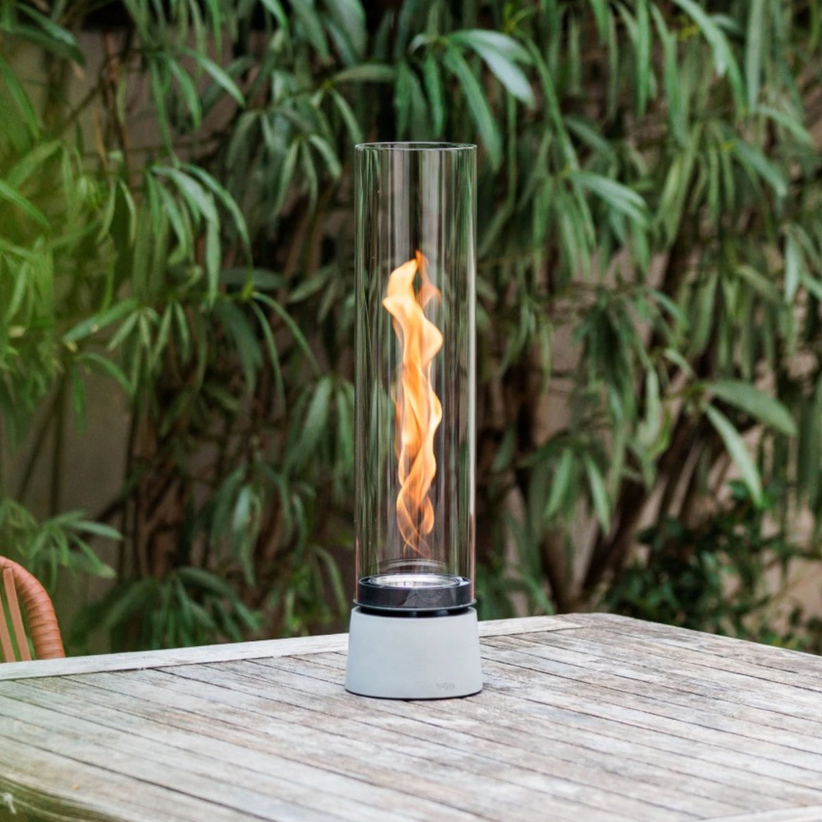 A tall cylindrical glass outdoor flame lantern with a realistic orange flame, set on a weathered wooden table with a green garden background.